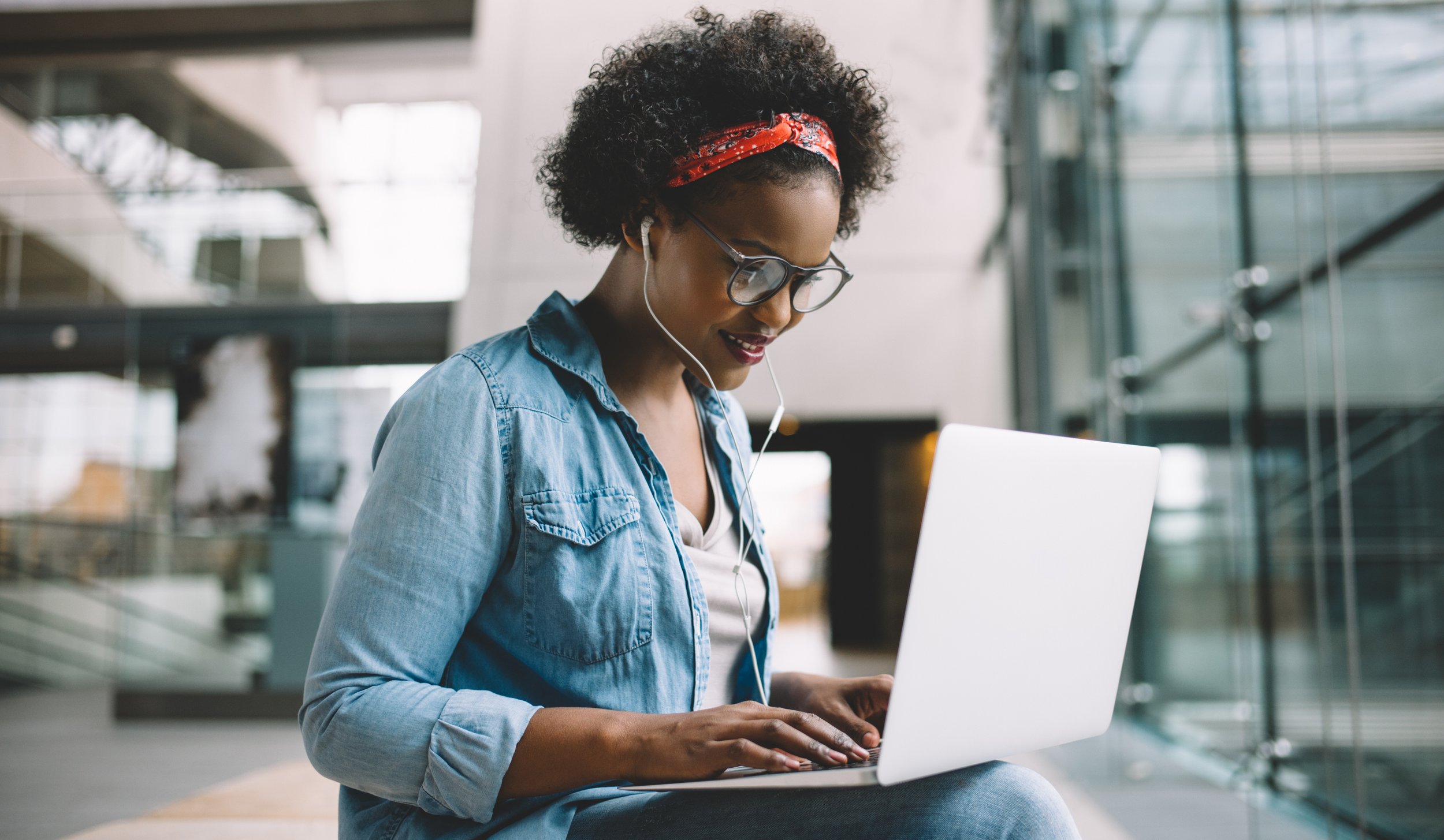 A woman with curly hair, glasses, and a red headband sitting indoors with a laptop, listening to music with earphones.