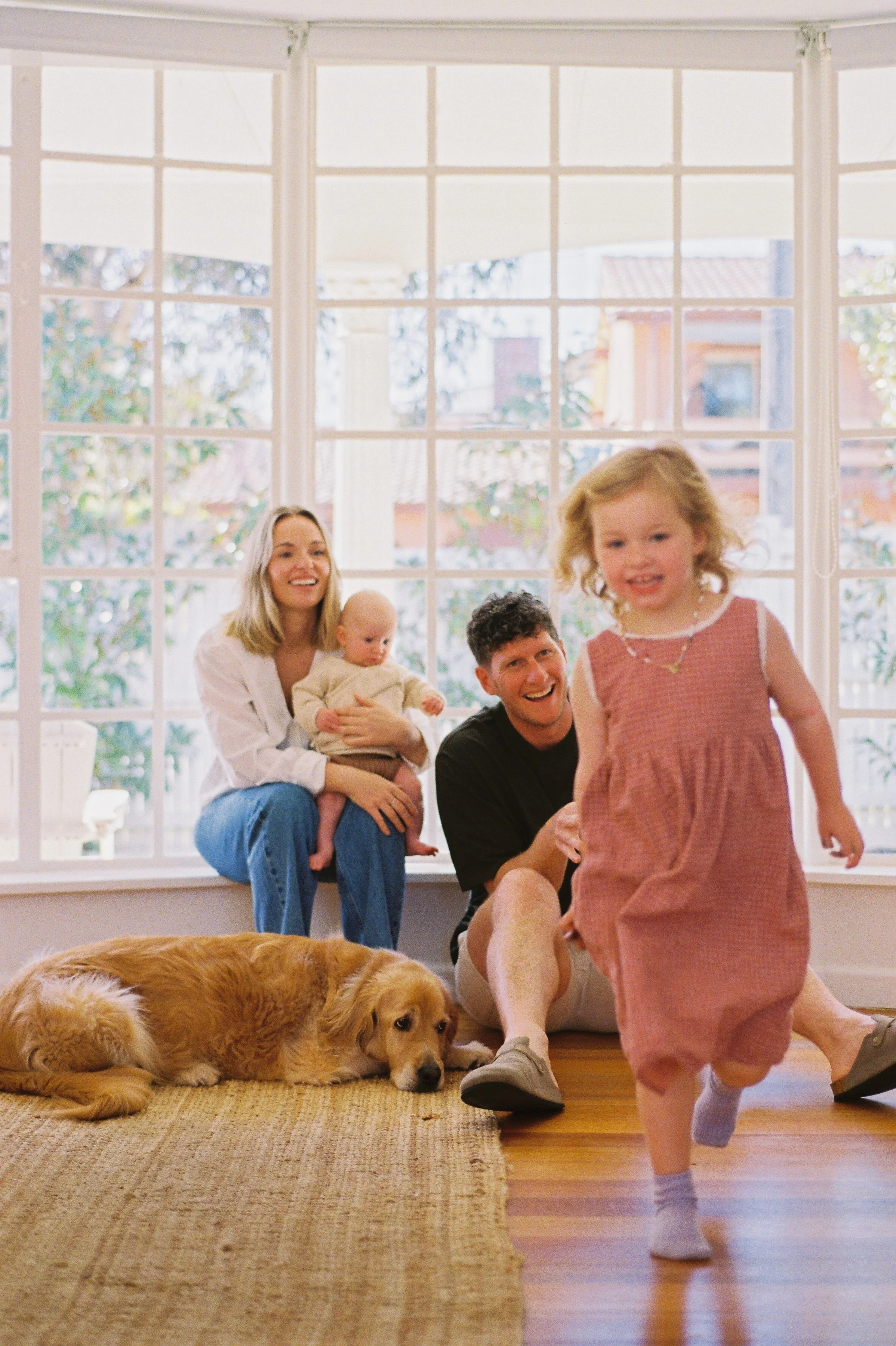 A family of four and their dog enjoying time together in a bright living room with large windows. The father is sitting on the floor, the mother sitting on the window sill holding a baby, a young girl is running toward the camera, and a golden retriever is lying on a rug.
