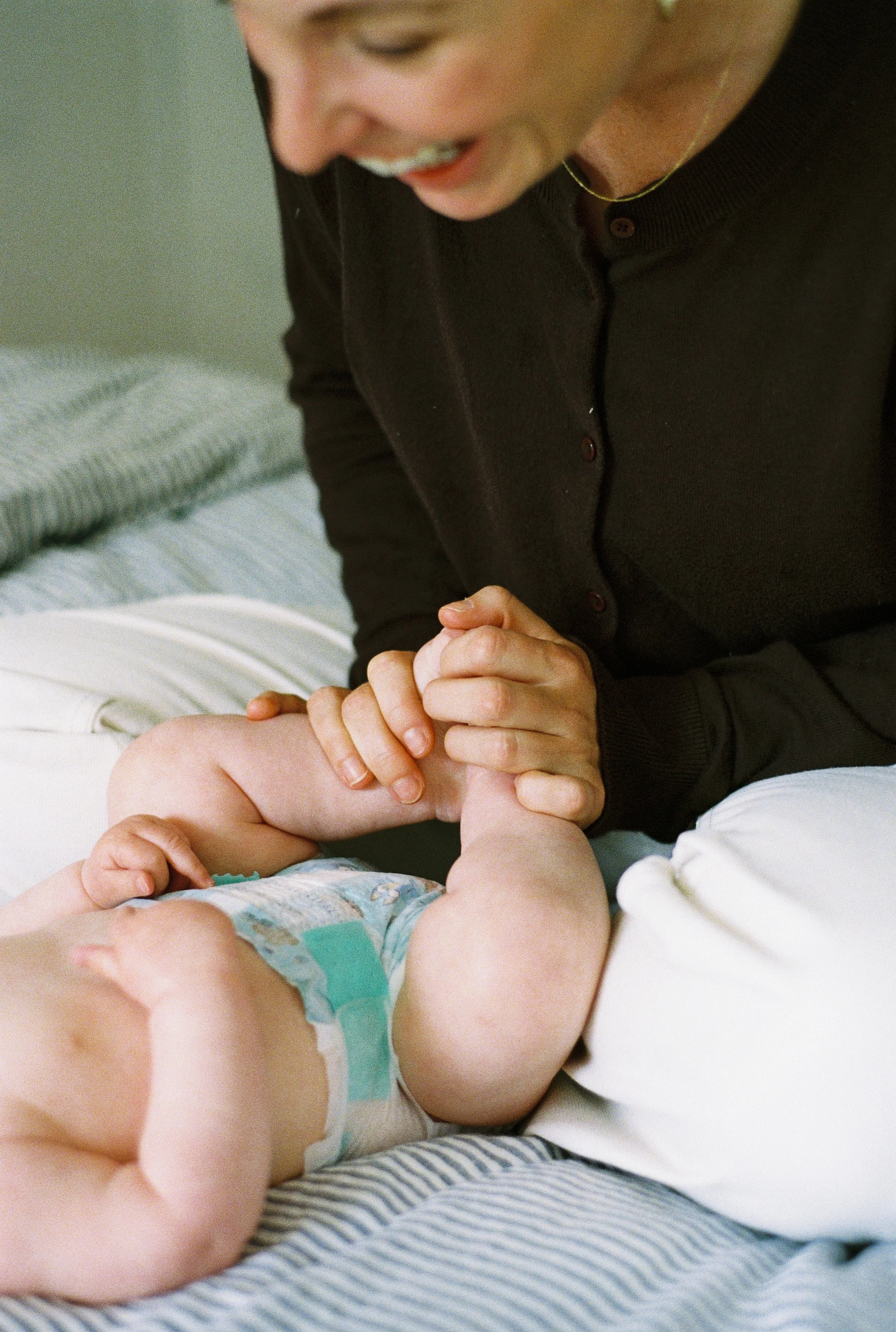 A woman playing with a baby on a bed, holding the baby's foot and smiling.