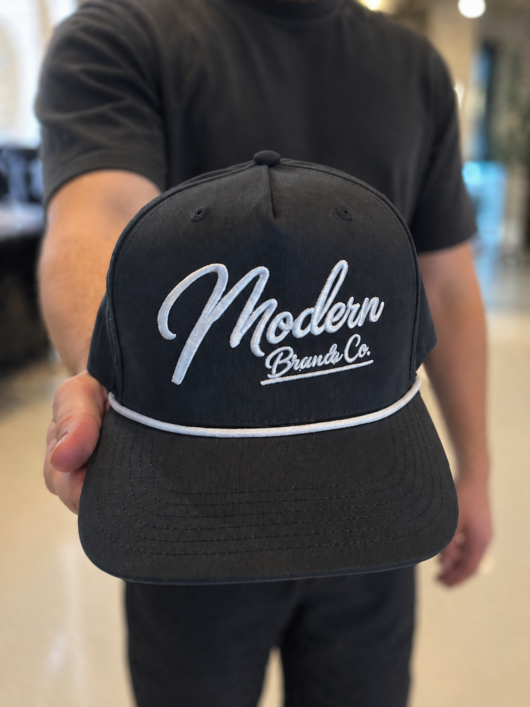 Person holding a black baseball cap with white embroidered text reading 'Modern Brands Co.'.