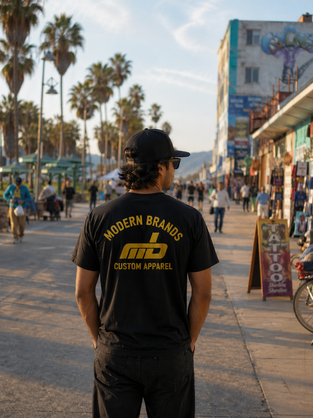 A young man wearing a black t-shirt with yellow letters that read "Modern Brands Custom Apparel" and a black baseball cap stands on a busy outdoor walkway. 