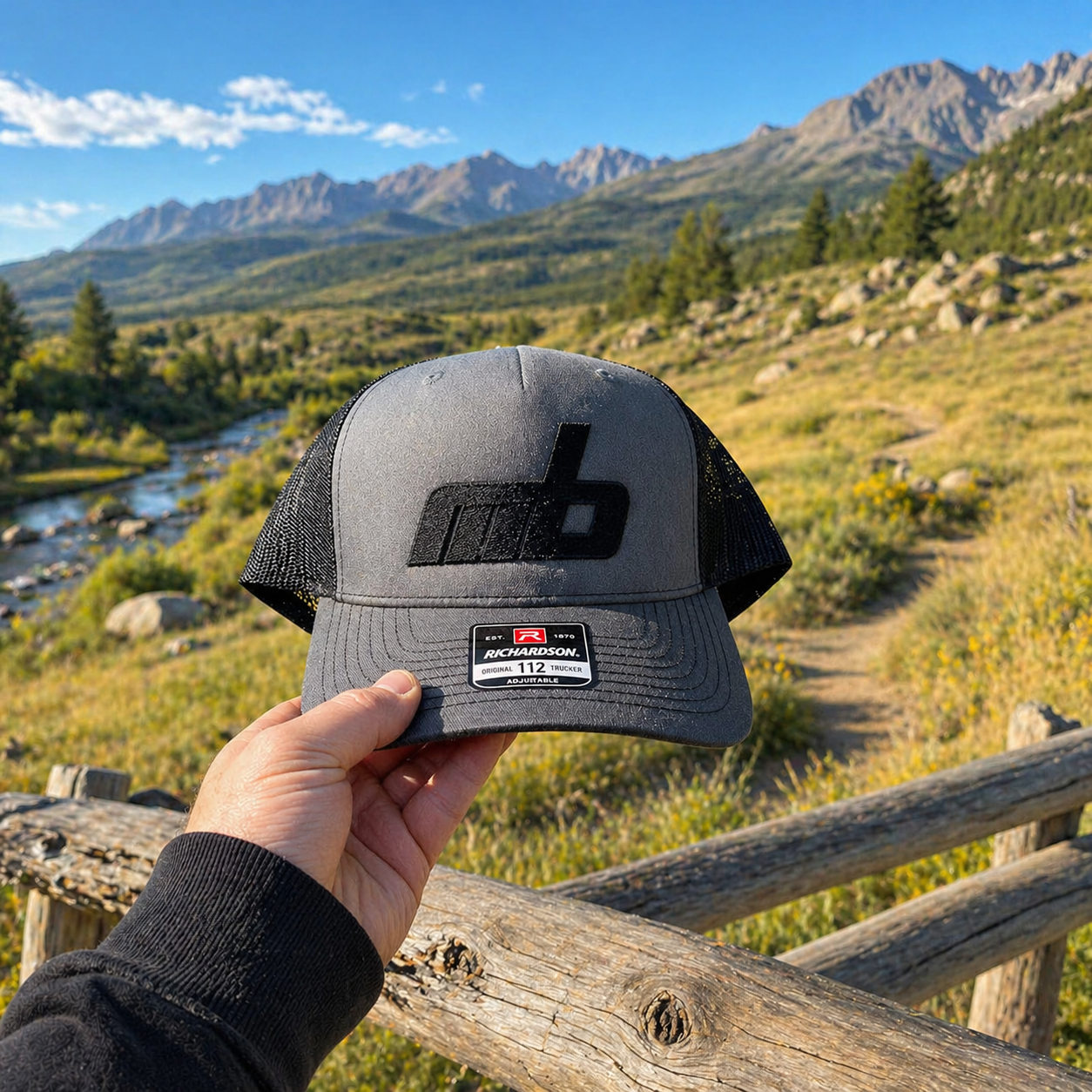 A hand holding a Richardson trucker cap with a mountain and river landscape in the background, under a blue sky with a few clouds.
