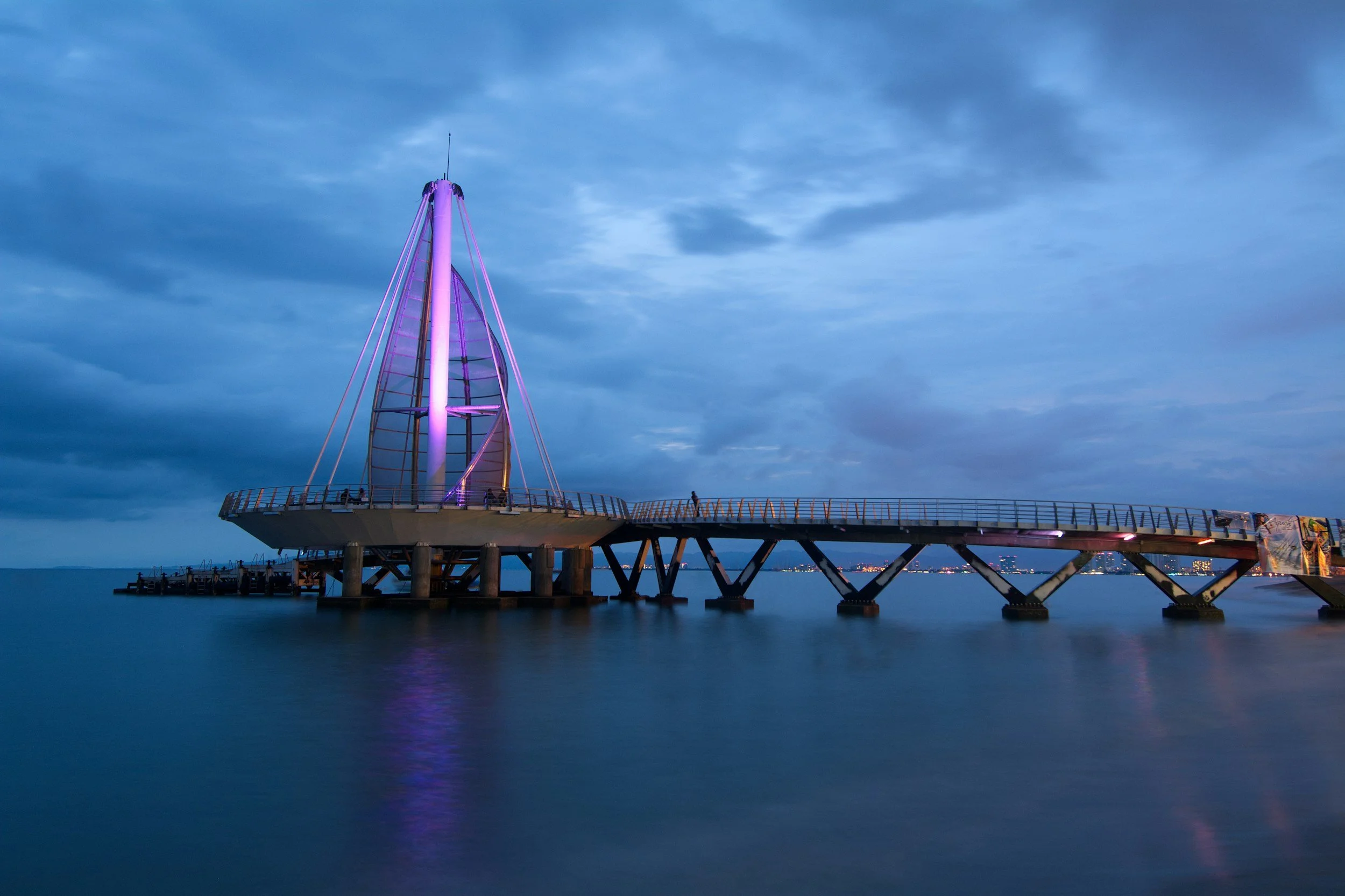 Pier with a modern, illuminated structure extending over a body of water, under a cloudy evening sky.