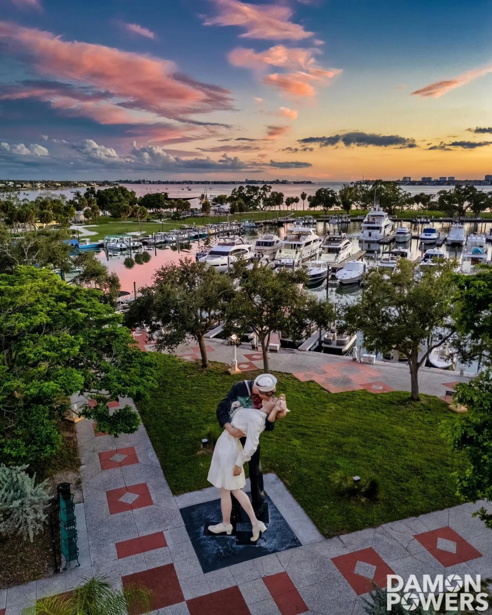 A statue of a man and woman kissing, located on a paved walkway with red and gray tiles, near trees and green grass, overlooking a marina with boats and water at sunset.