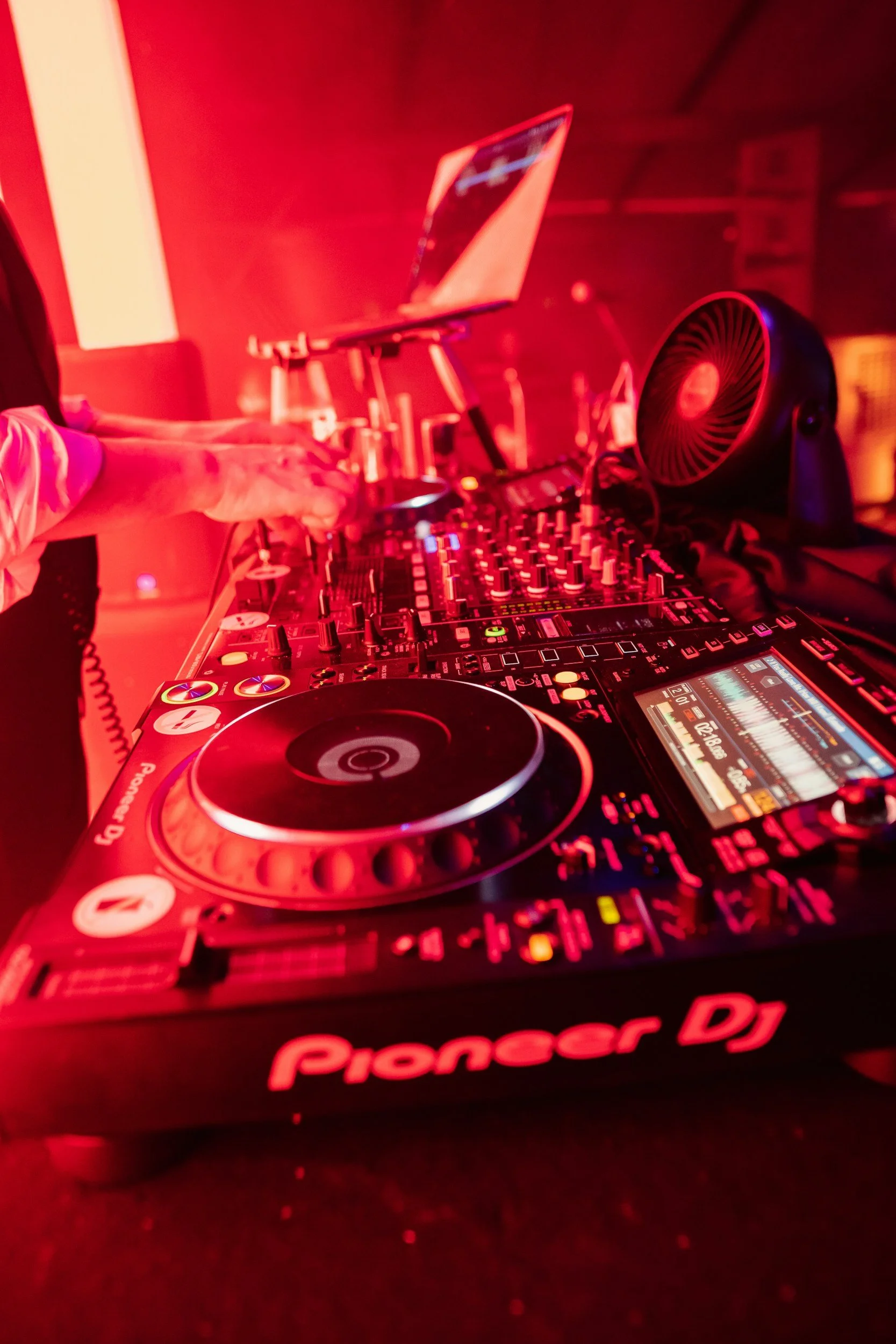 Close-up of a DJ controller with a person's hands adjusting controls, illuminated by red lighting, with a laptop in the background.