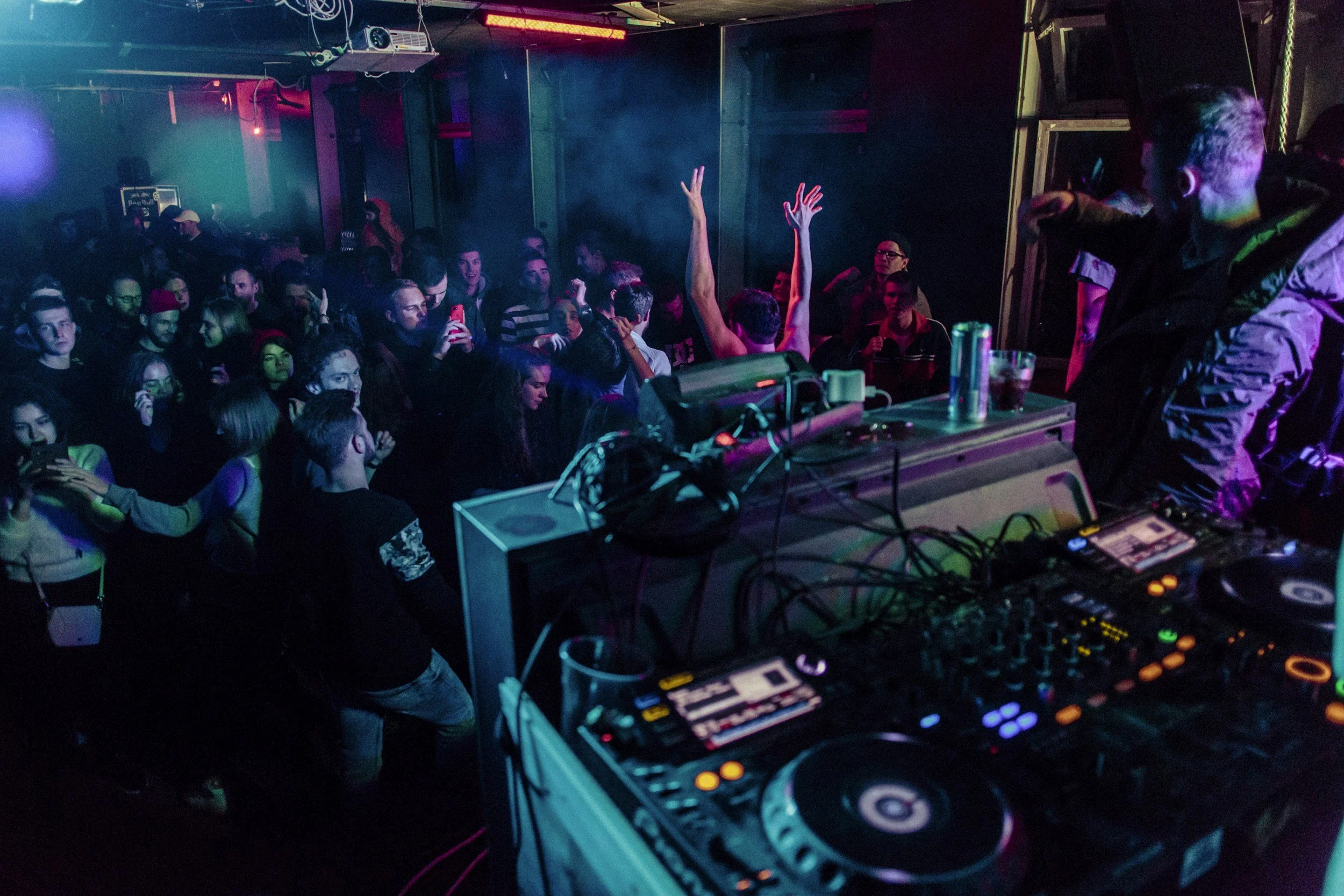 People dancing and socializing in a nightclub with DJ equipment and colorful lighting.