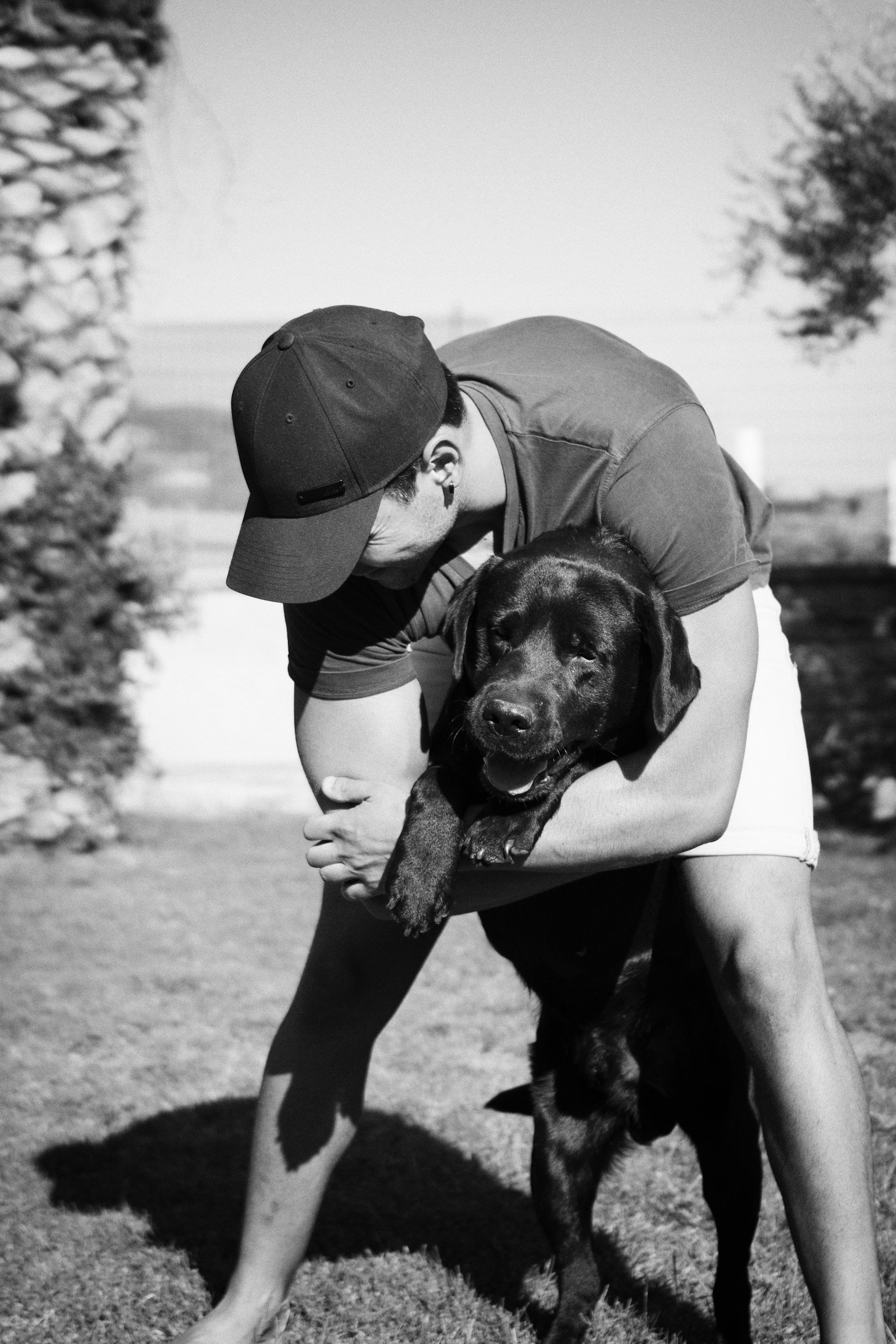 A person hugging a large black and brown dog outdoors on grass, with trees in the background, in black and white.