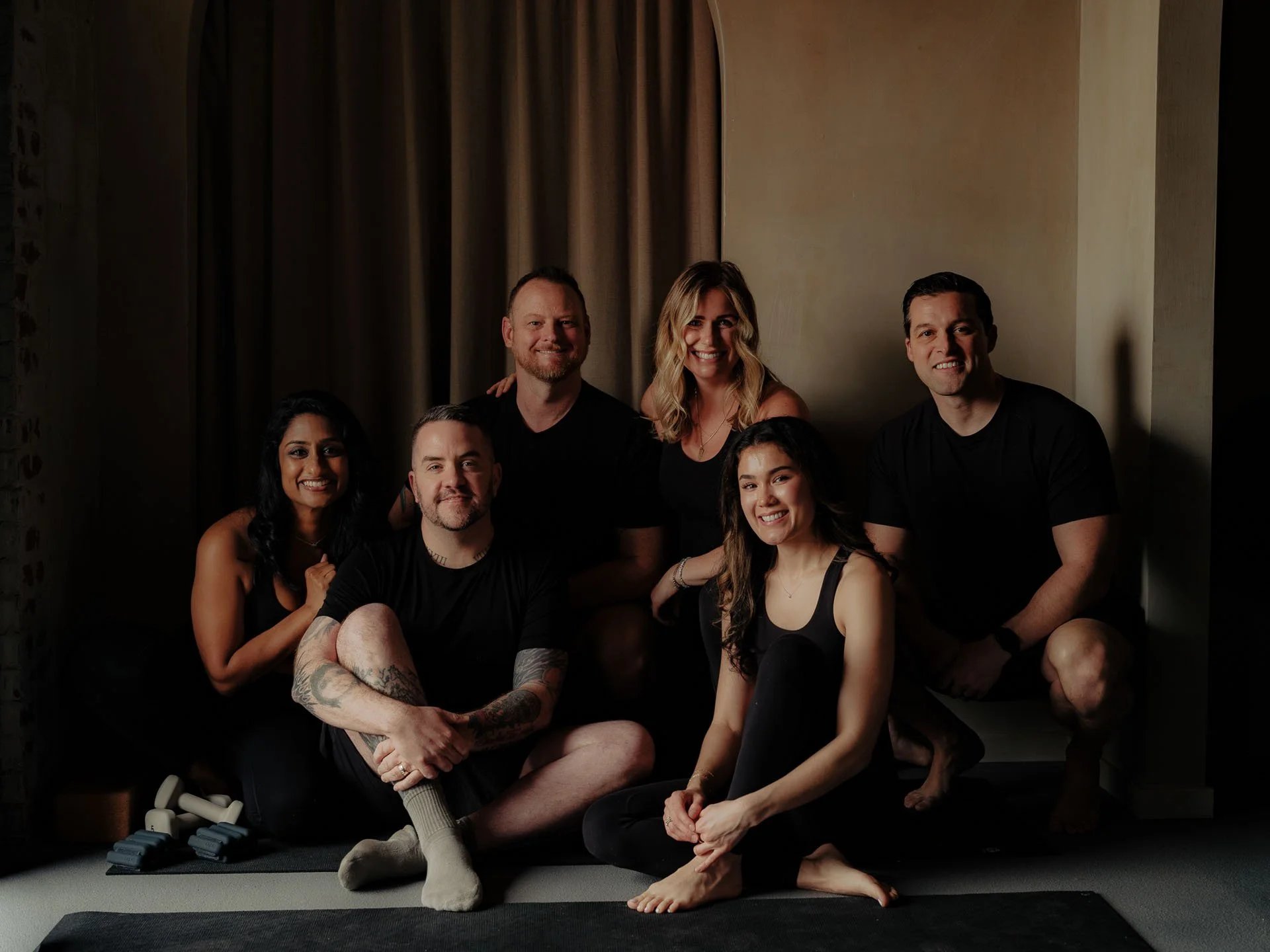 Group of six people posing indoors, sitting and kneeling on the floor, smiling at the camera, with a beige wall and curtains in the background.