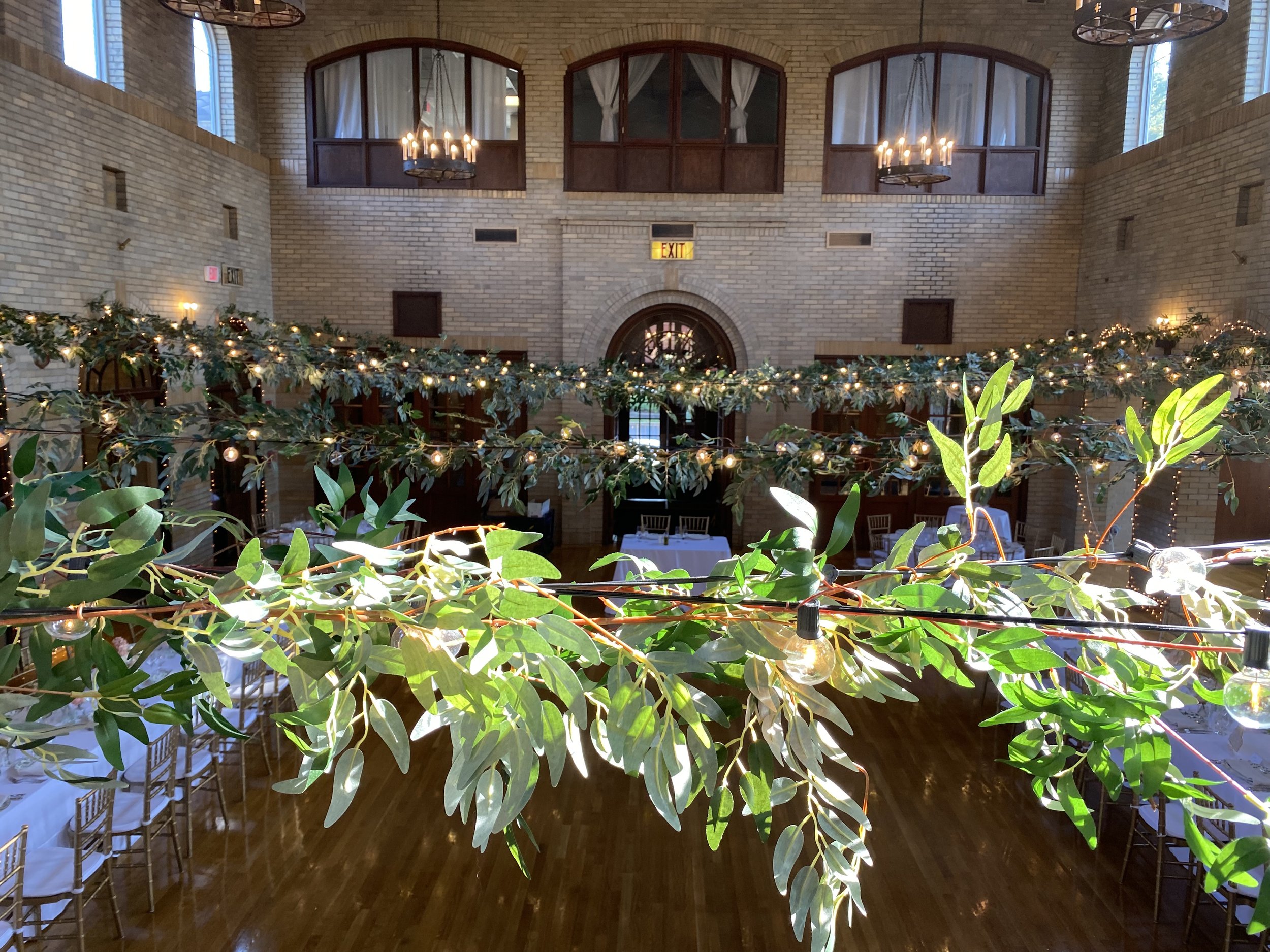 Decorative string lights and green foliage hanging in a banquet hall with brick walls, high windows, and chandeliers.
