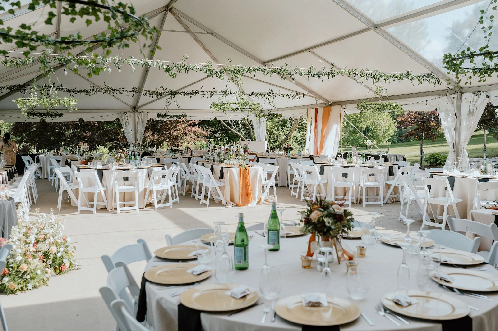 Decorated outdoor wedding or event reception under a white tent with round tables, white chairs, floral centerpieces, and greenery hanging from the ceiling, with a lush green landscape outside.