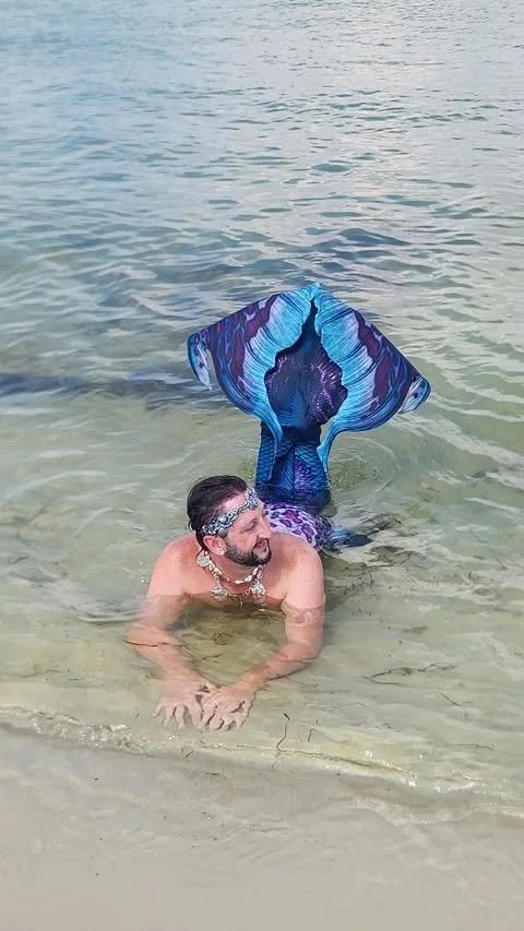 Man with a mermaid tail in shallow water at the beach, wearing sunglasses and a shell necklace.