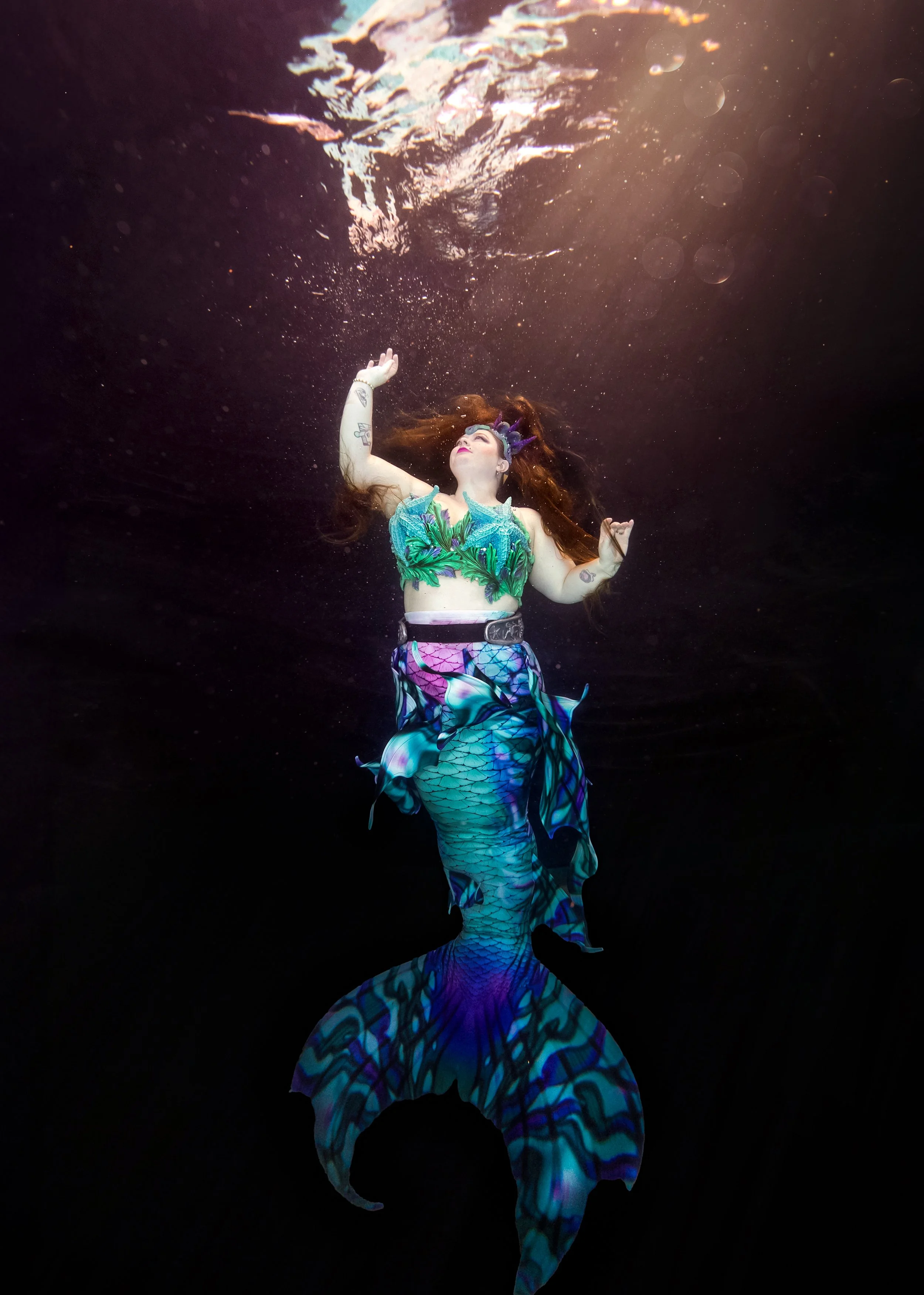A woman with long red hair and tattoos is underwater wearing a mermaid costume, with a green leafy top and a multicolored tail, gazing upwards with her arms raised.