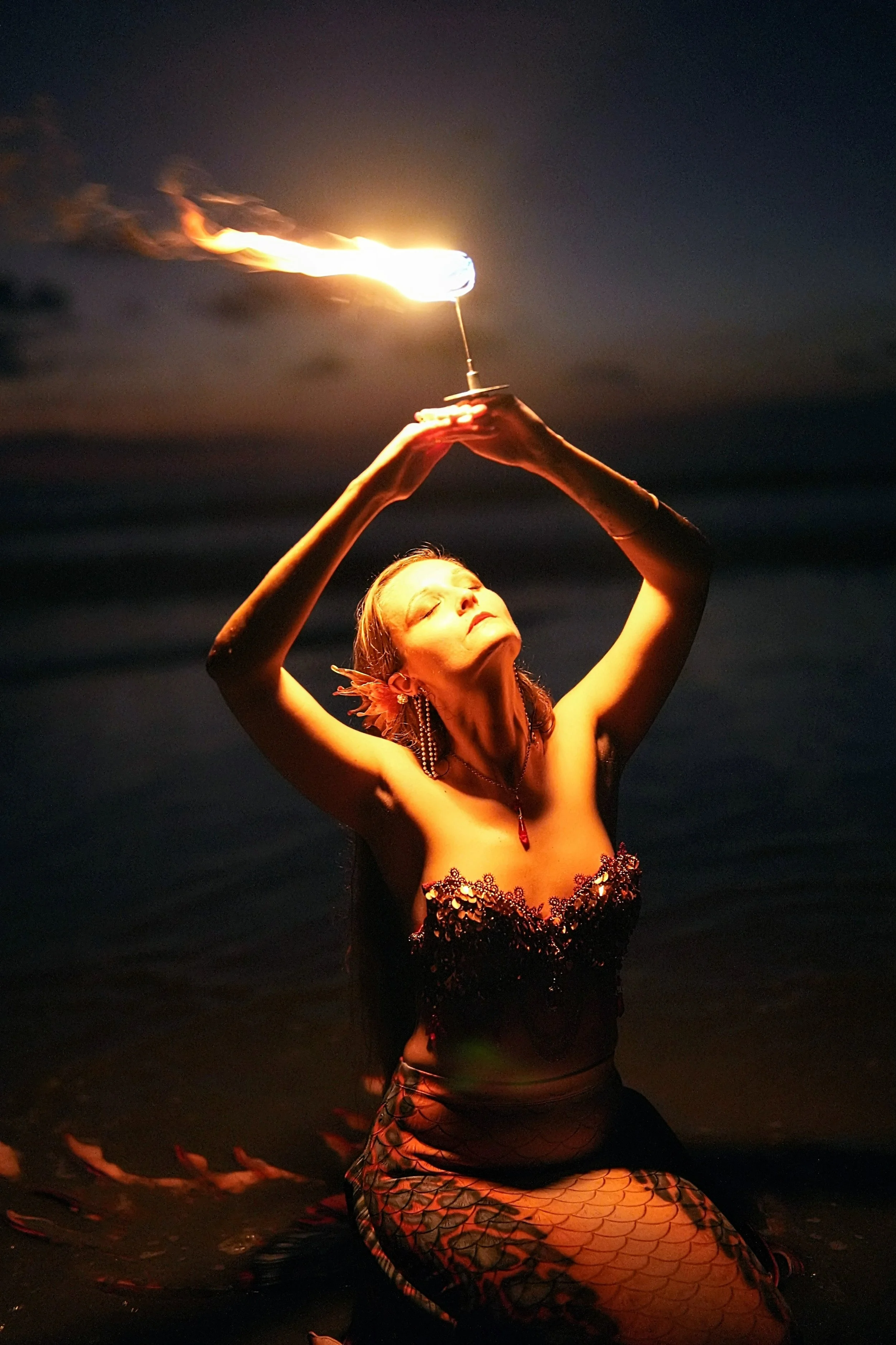 A woman in a mermaid costume performing a fire dance at night on the beach, holding a flaming torch above her head.