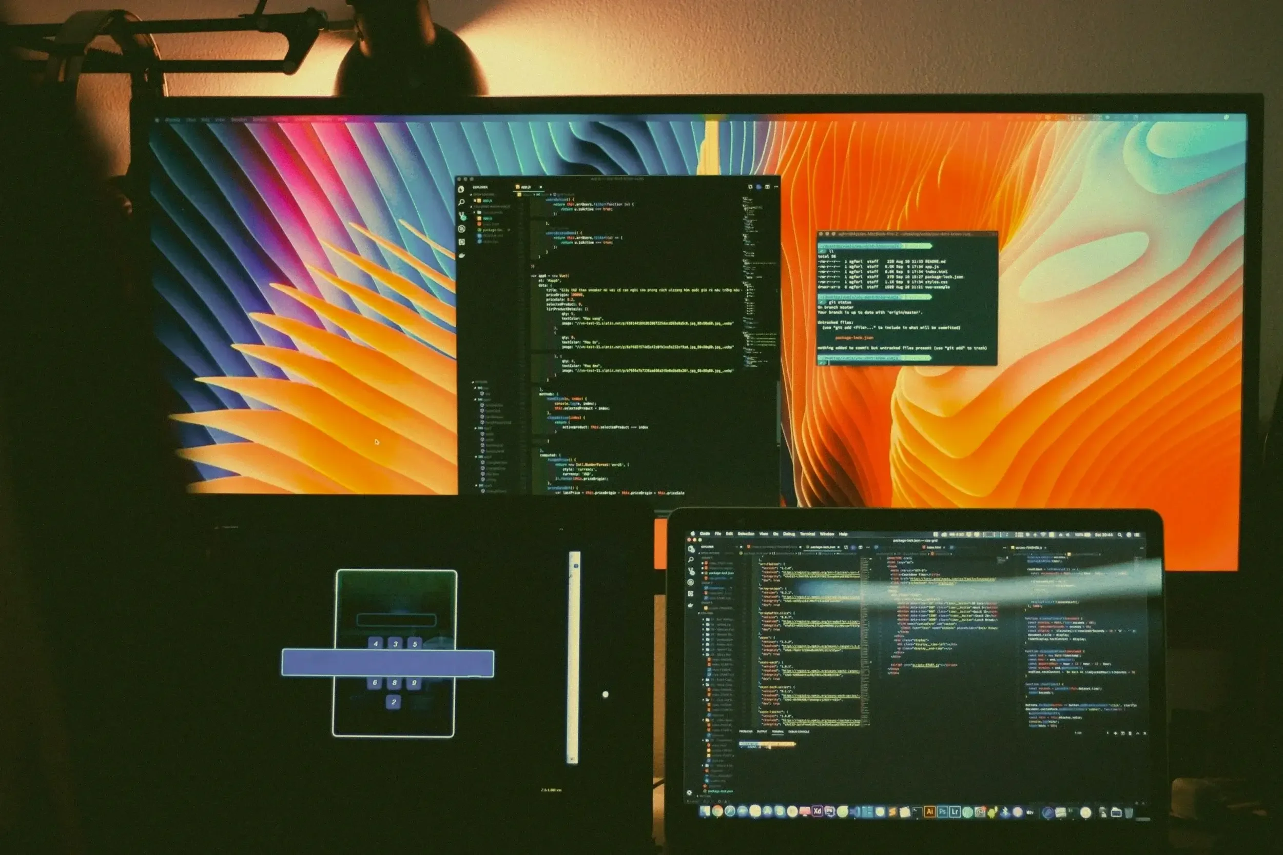 A workspace with a large screen displaying colorful abstract background and code editors, and a laptop showing code, in a dimly lit room.