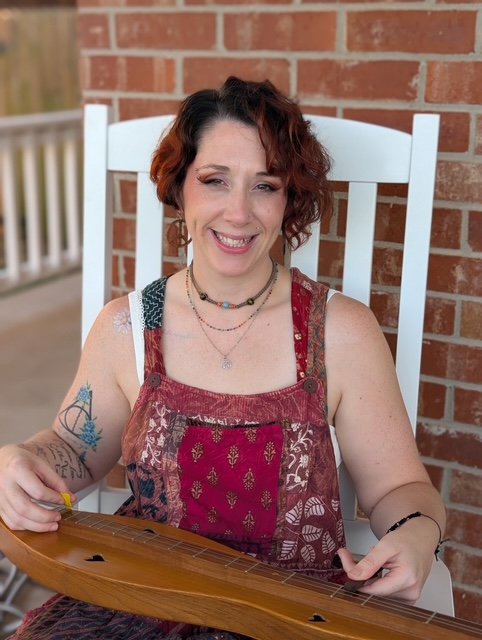 A portrait of a licensed professional counselor in Oklahoma City smiling warmly, sitting outside  holding a dulcimer.