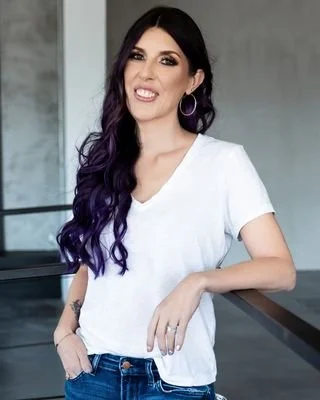 A portrait of a licensed clinican social worker in Oklahoma City smiling with long dark hair with purple highlights, wearing a white t-shirt and hoop earrings, standing indoors with a modern background.