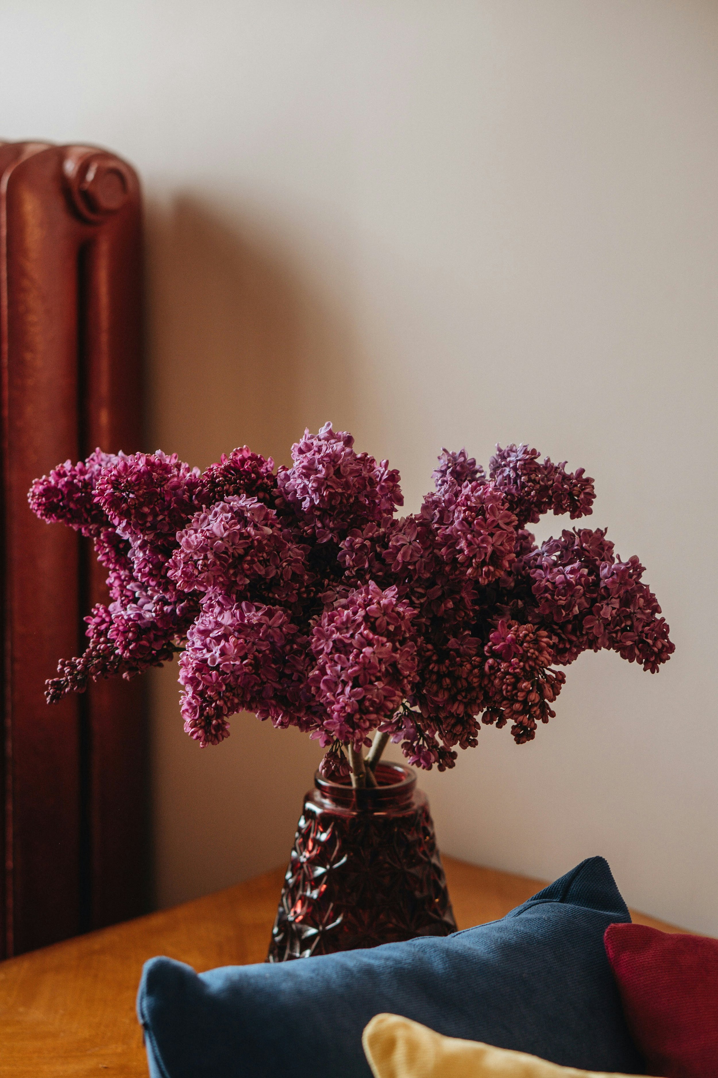 A bouquet of purple lilacs in a textured dark red vase on a wooden surface, with colorful pillows in the foreground and a plain light-colored wall in the background.