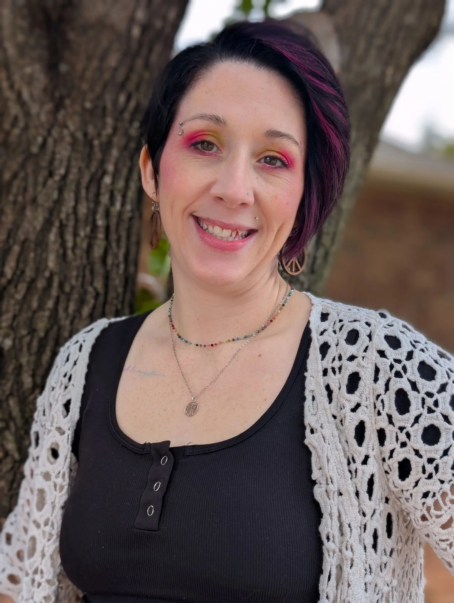 A woman with short dark hair and purple highlights is smiling outdoors in front of a tree. She is wearing colorful eye makeup, earrings, layered necklaces, a black top, and a white crocheted cardigan.