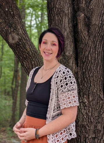 A woman with short dark hair, pink highlights, and a warm smile standing outdoors against a tree, wearing a black top, a white crocheted cardigan, and brown pants.