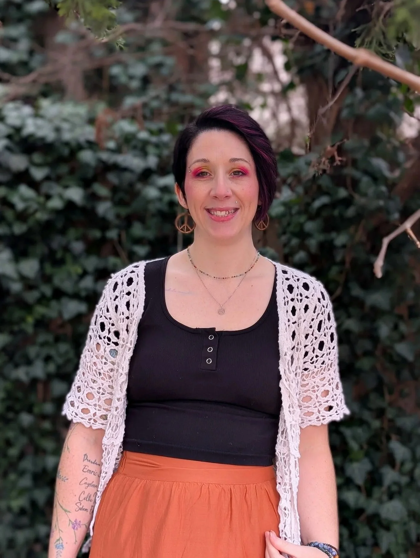 A woman with short dark hair and pink makeup, smiling in front of lush green foliage, wearing a black top, a white crocheted cardigan, and an orange skirt, with jewelry and tattoos on her arms.