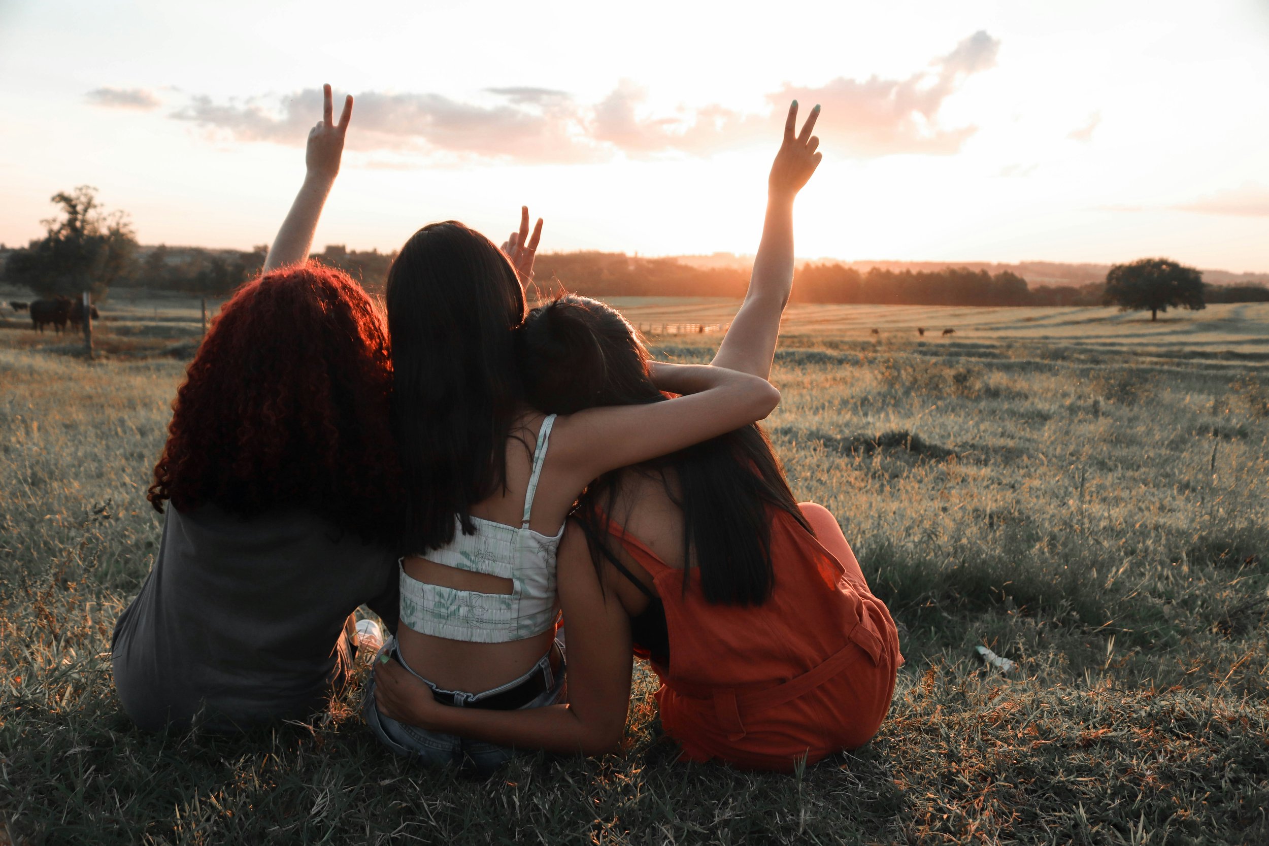 Three women sitting on the grass, looking at the sunset, with two of them making peace signs with their hands.