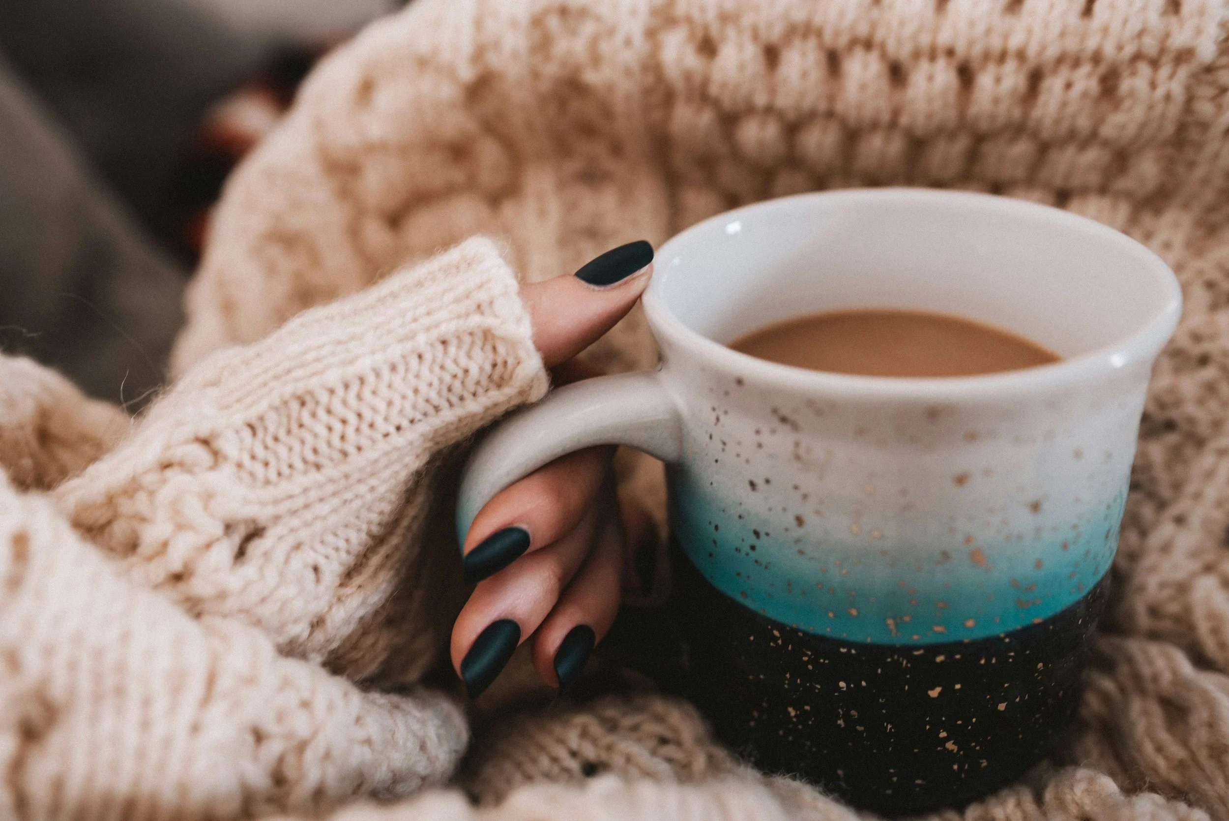 Close-up of a person holding a coffee mug with hands in cozy beige cable-knit mittens, surrounded by chunky knit beige sweaters.