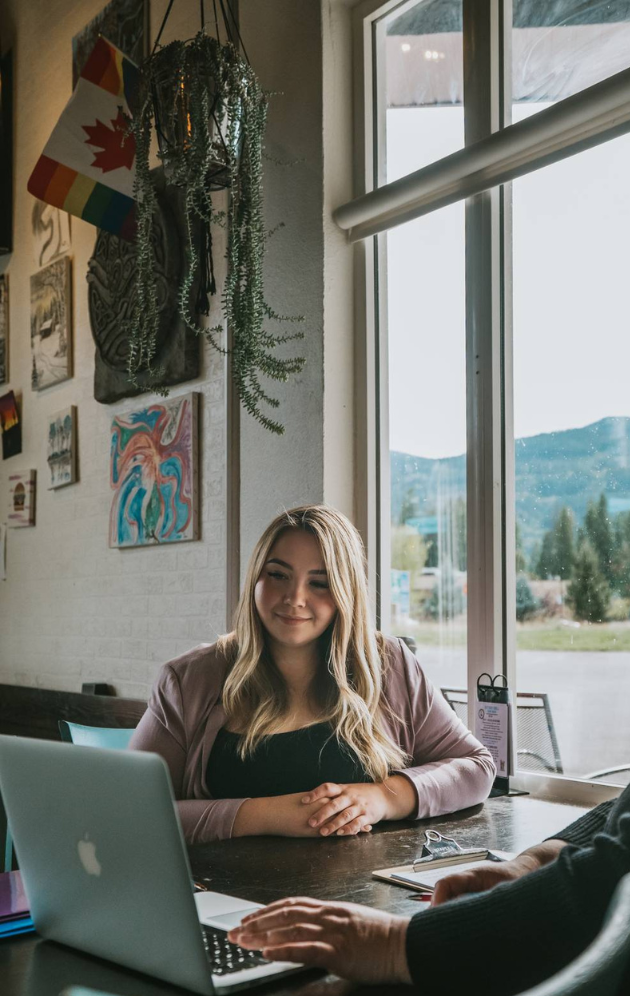 A young woman with blonde hair sitting at a table, smiling while using a laptop in a cozy cafe with artwork and plants on the wall, large windows showing a scenic view outside.