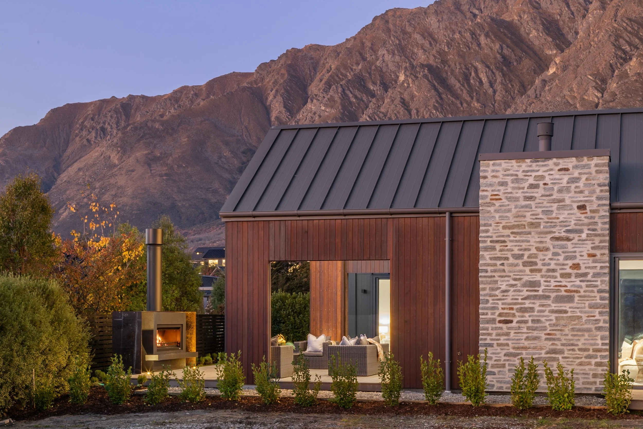 New build with brick chimney and metal roof against mountain backdrop at dusk — Jacks Point, Queenstown