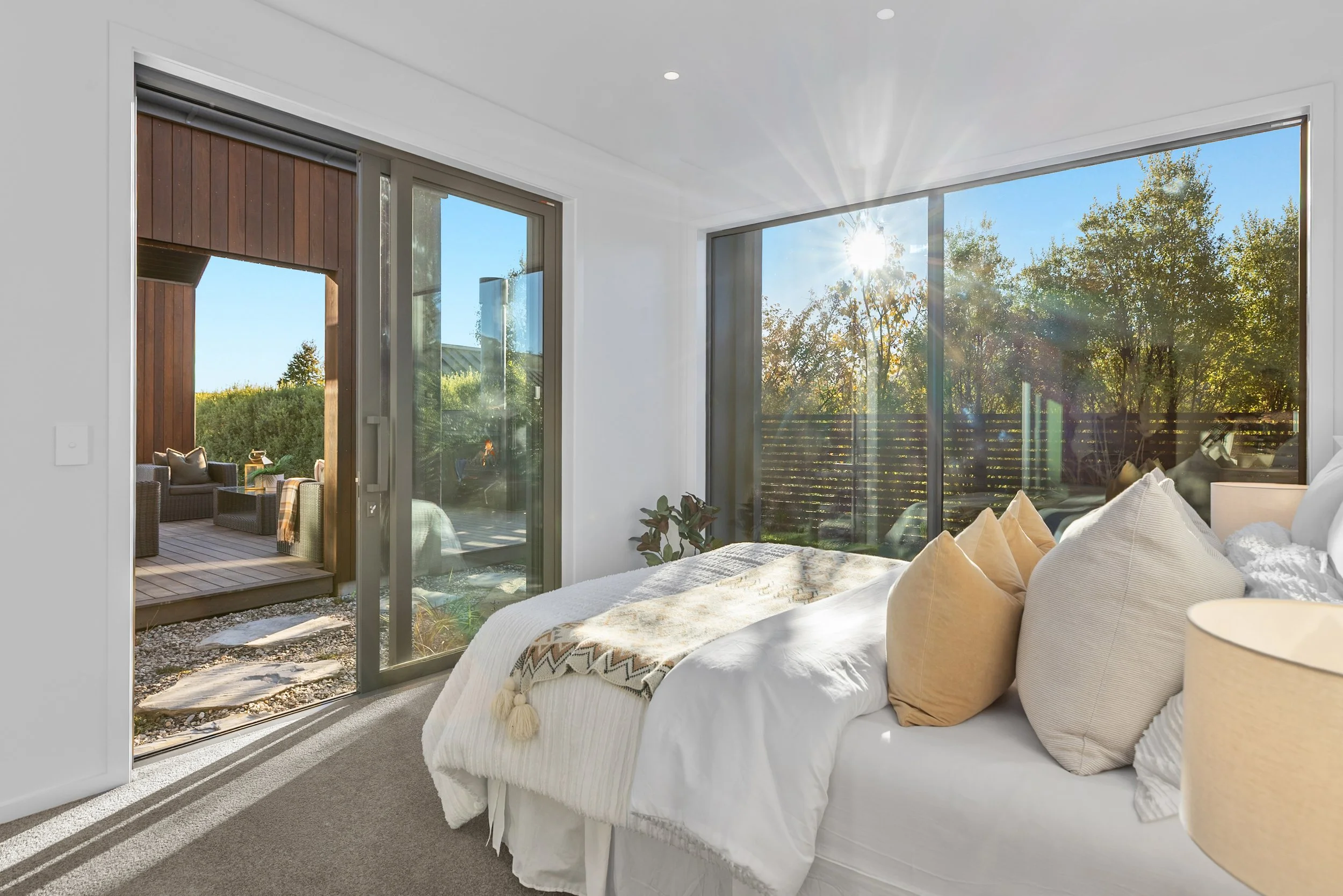 New build bedroom with sliding doors to patio and wicker chairs, sunlit trees outside — Jacks Point, Queenstown