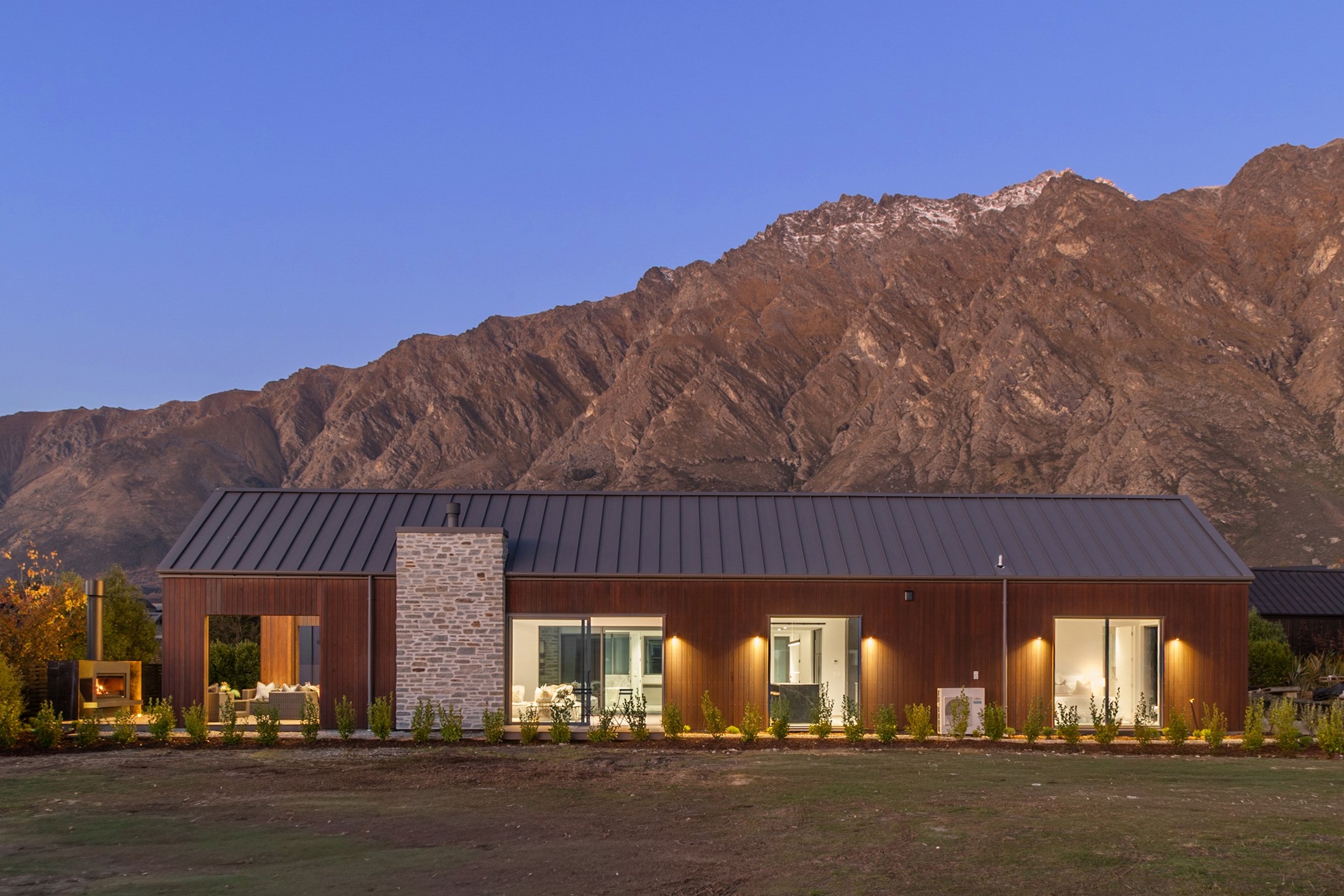 New build with large windows and outdoor lighting design against mountain backdrop at dusk — Jacks Point, Queenstown