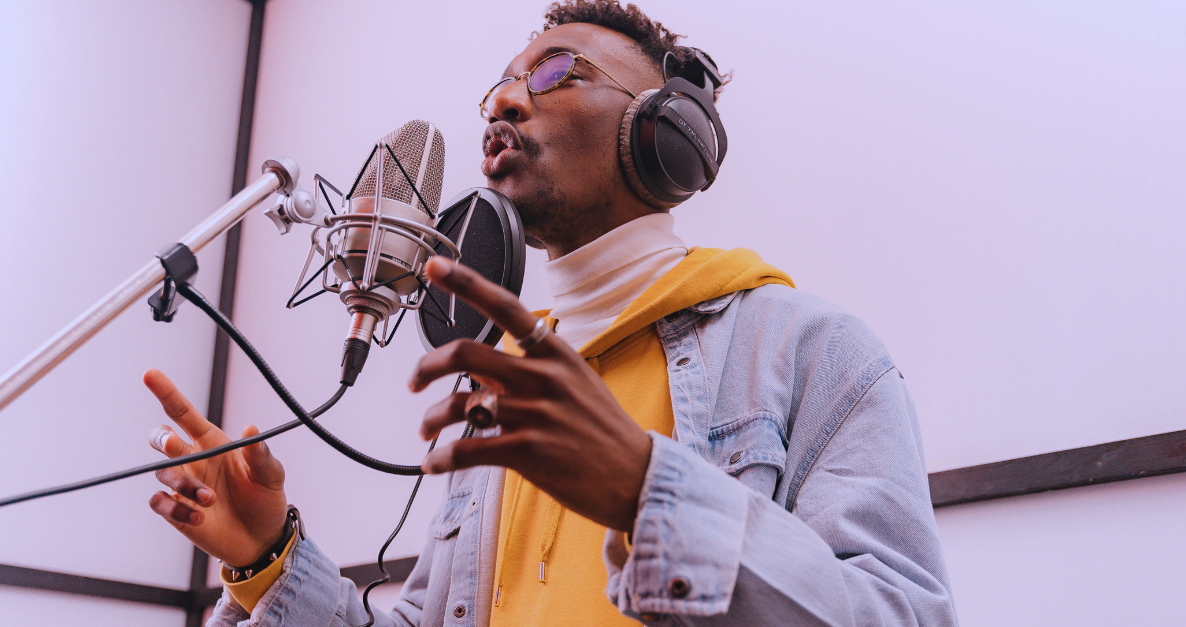 A man singing into a professional microphone in a recording studio, wearing headphones and glasses, with a purple background.