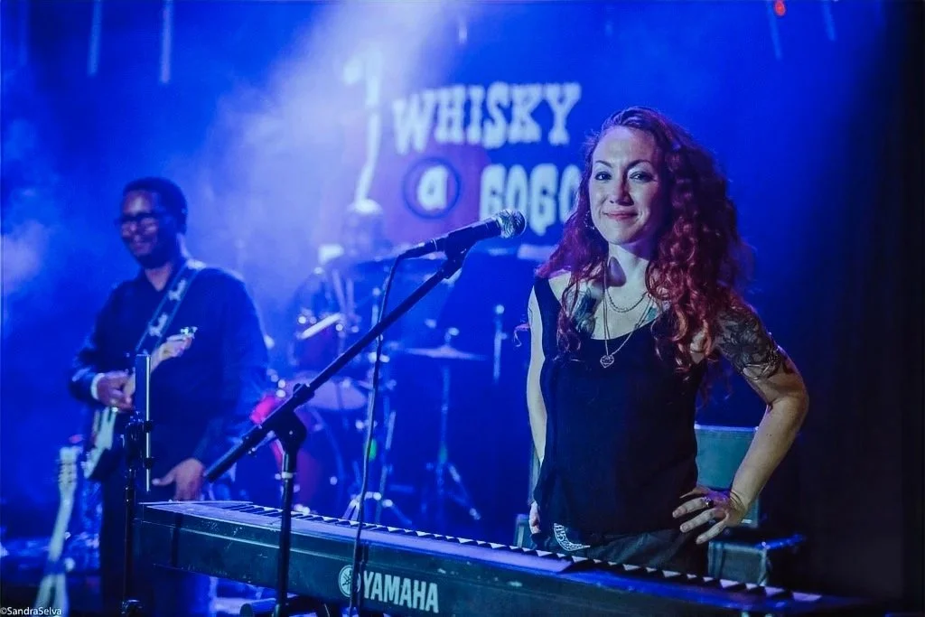 A woman with curly red hair standing behind a keyboard on stage, smiling, with a band in the background at a music venue called Whiskey A Go Go.