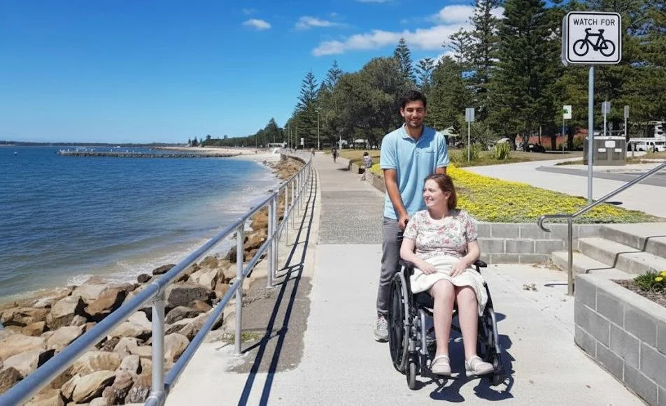 A young man and a woman in a wheelchair on a seaside promenade on a sunny day, with a 'Watch for Bicycles' sign nearby.