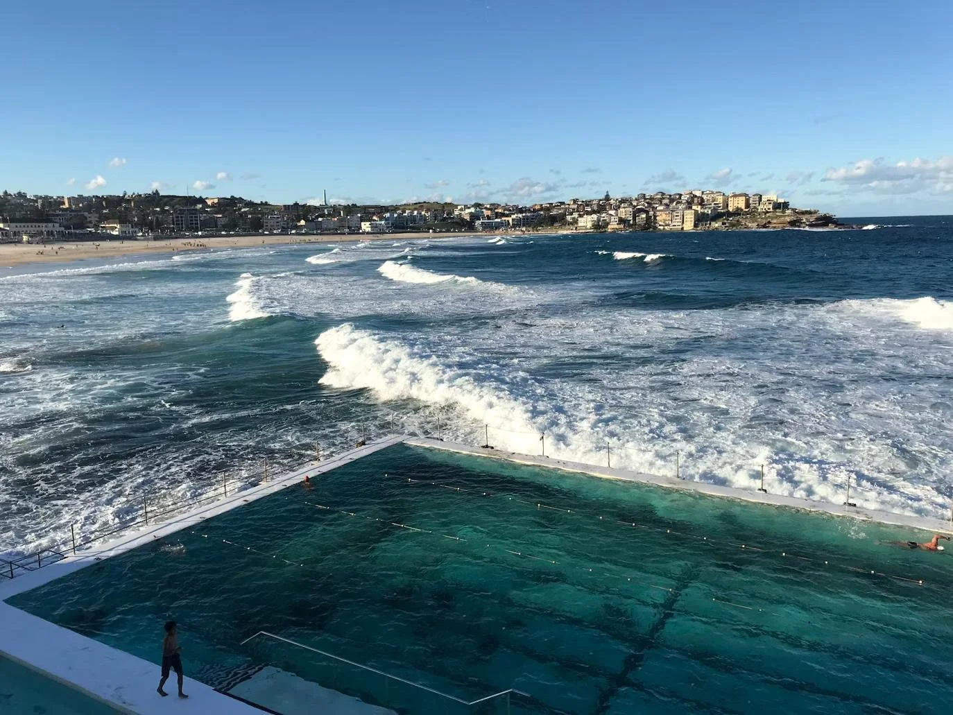 A swimming pool on a boat, with ocean waves and a distant shoreline and buildings in the background under a blue sky.