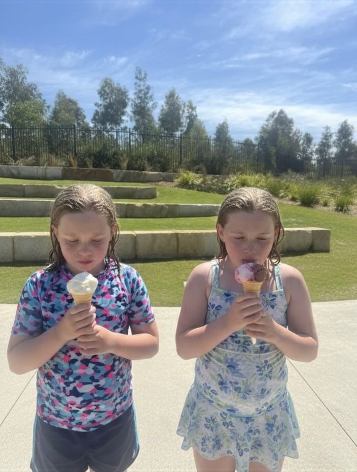 Two young girls standing outdoors on a sunny day, each holding an ice cream cone and wearing summer clothes, with green lawn and trees in the background.