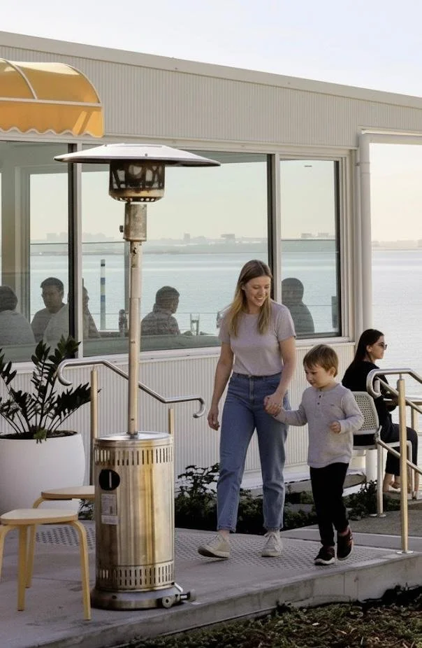 A woman and boy walk outside on a patio near a large window by a body of water, with other people visible through the window. There is a patio heater and plant nearby.