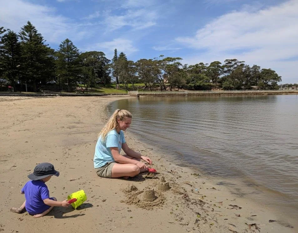 A woman and a small child playing on a sandy beach near a body of water. The woman is sitting on the sand, building a sandcastle with a red shovel. The child, wearing a purple shirt and a wide-brimmed hat, is sitting nearby with a yellow toy and a re