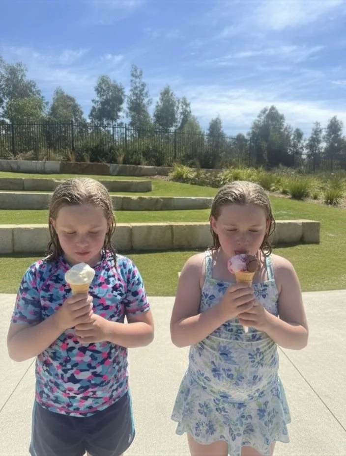 Two young girls standing outdoors on a sunny day, each holding a cone with ice cream, with a background of grass, steps, and trees.
