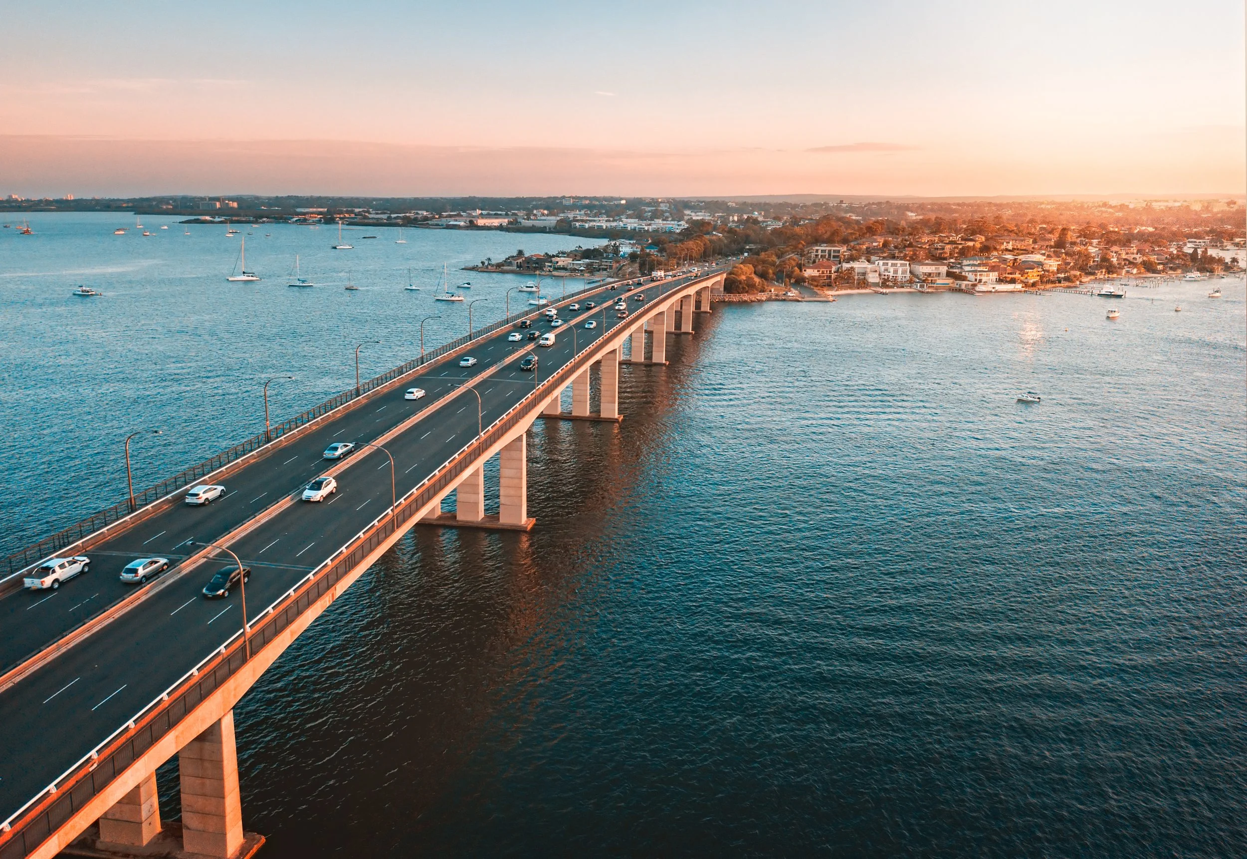 Aerial view of a bridge extending over a body of water with boats, leading toward a cityscape at sunset.