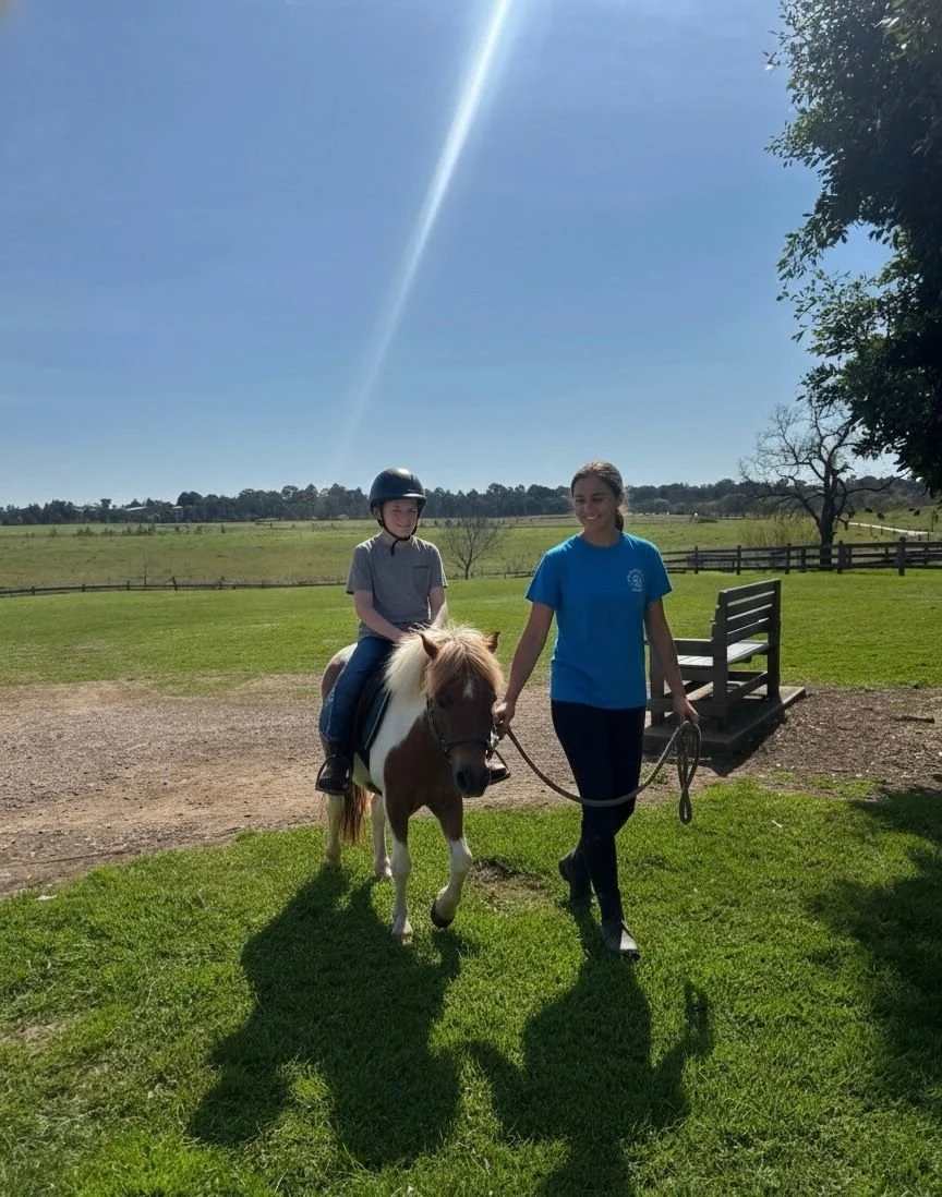 A young boy riding a small horse or pony, guided by a woman holding the reins, on a sunny day at a riding stable or farm.