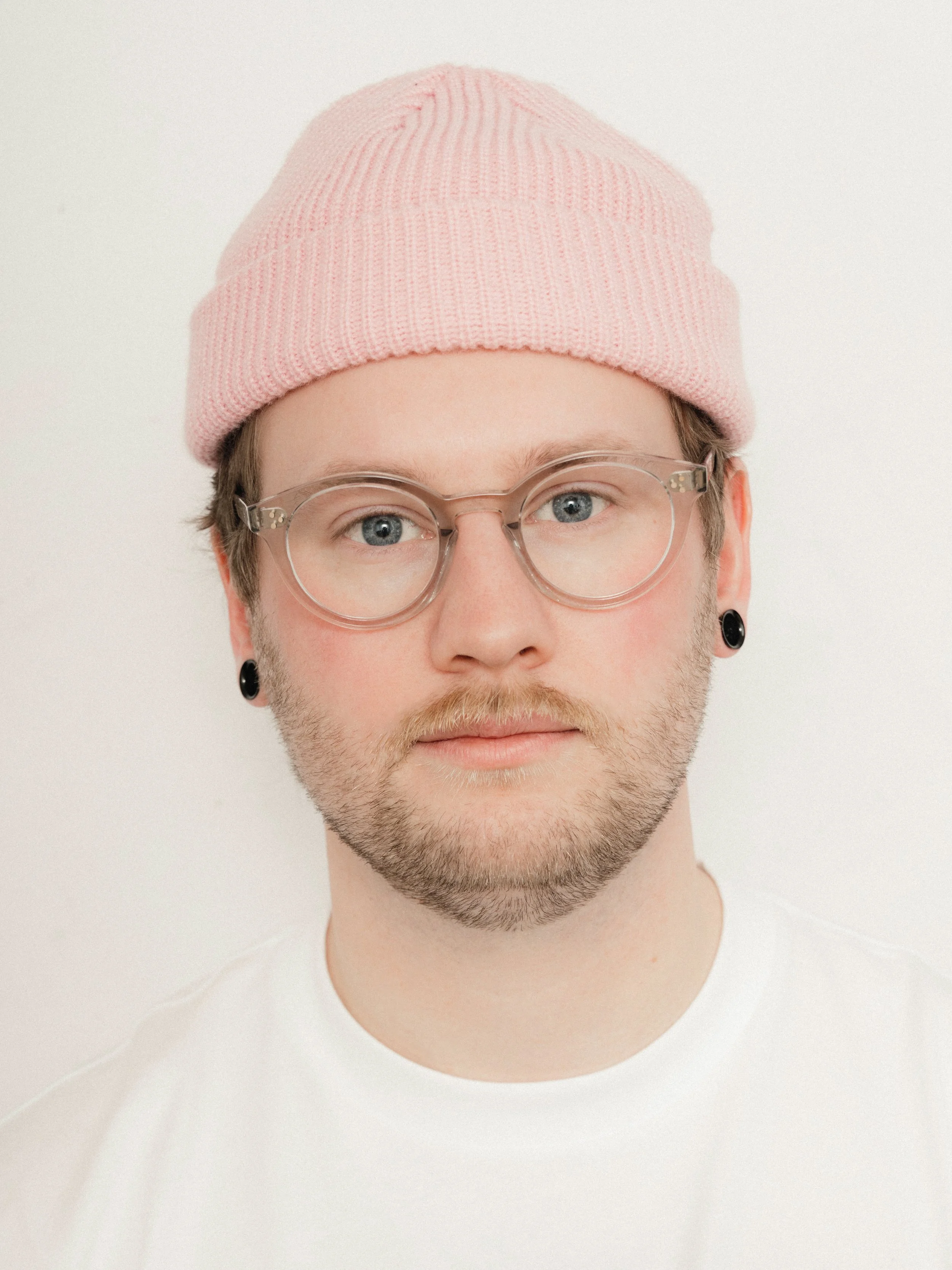 Porträt eines jungen Mannes mit Brille, pinkem Mütze, schwarzen Ohrsteckern und hellbraunem Bart vor weißem Hintergrund.