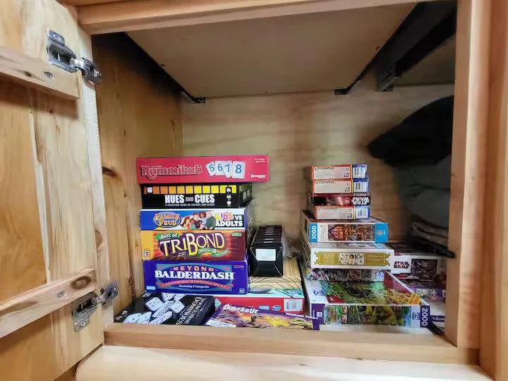 Inside a wooden cabinet, various board games and puzzles are stored, including titles like Rummikub, Hues and Cues, Candy Land, Tri Bond, Beyond Balderdash, and several others, with some stacked on top of each other.