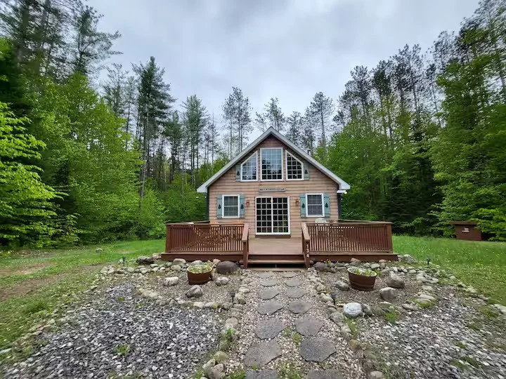 A cabin with large windows, a wooden deck, and a stone pathway leading to the front door, surrounded by green trees.
