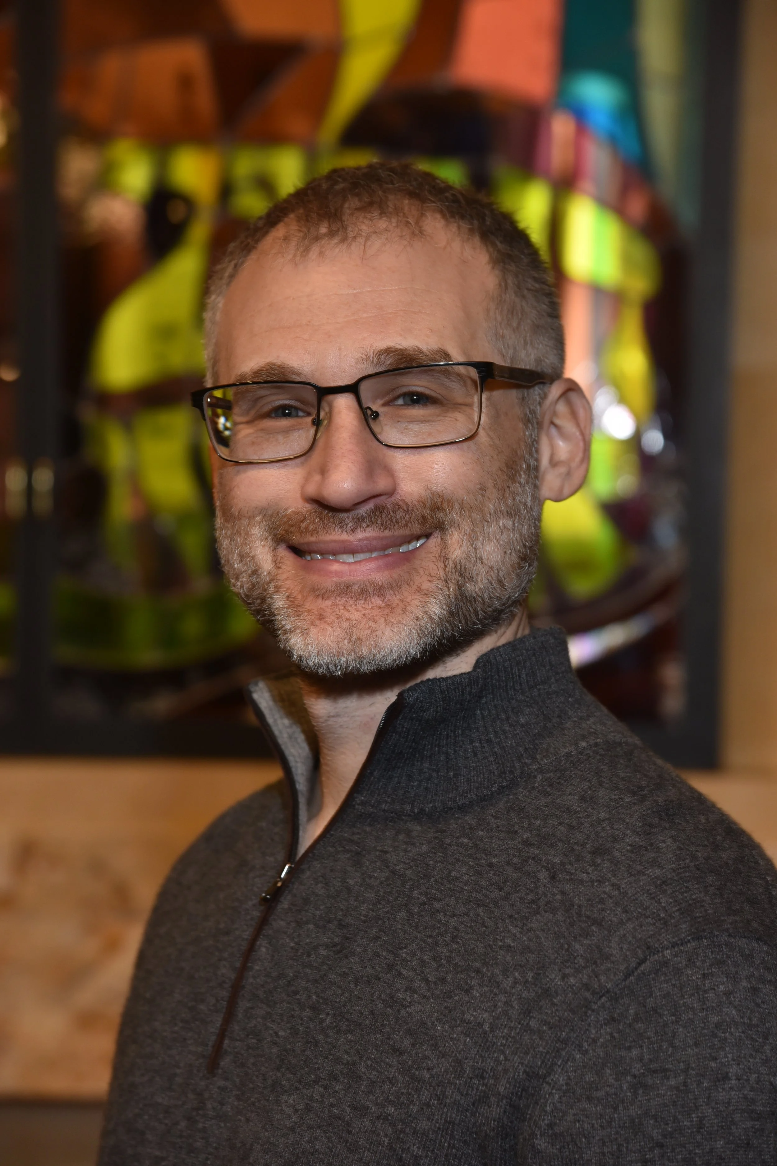 A smiling man with glasses and a beard wearing a gray quarter-zip sweater, standing in front of a colorful, abstract art piece.