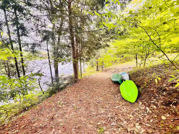 Two kayaks and a small boat resting on the ground beside a wooded trail near a pond.