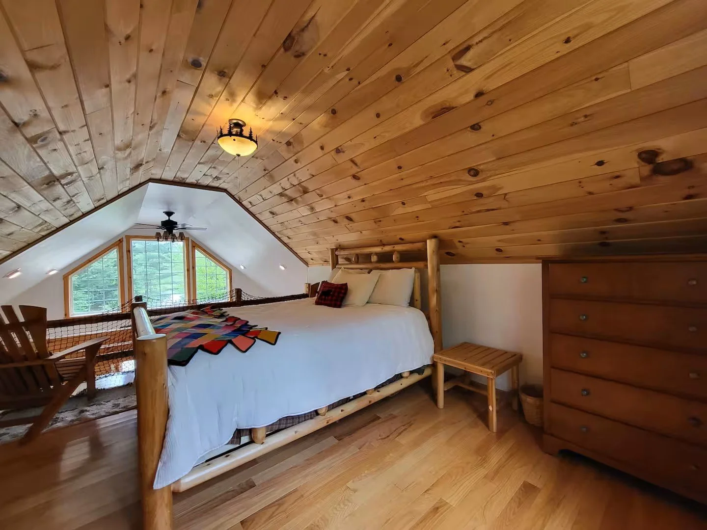 A cozy attic bedroom with a wooden sloped ceiling, a bed with white bedding, colorful quilt, and pillows, a small wooden nightstand, and a large window with trees outside.