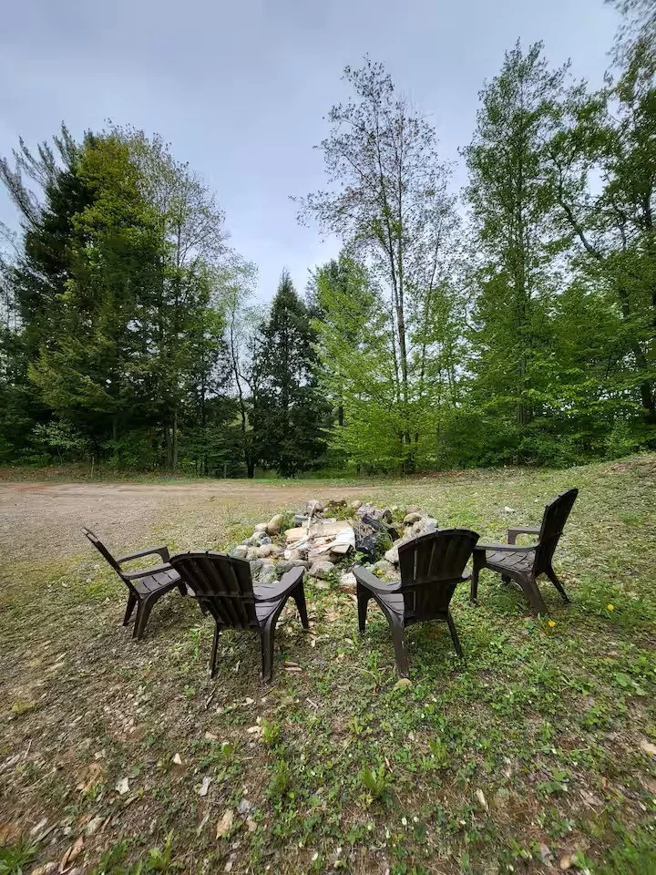 Four black outdoor chairs arranged in a circle around a fire pit made of rocks and wood, set on grassy ground with trees in the background under a cloudy sky.
