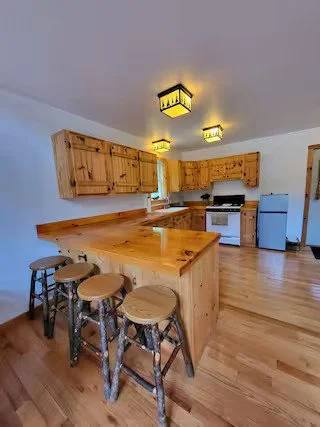 Kitchen with wooden cabinets, counter with stools, a stove, refrigerator, and hardwood floors.
