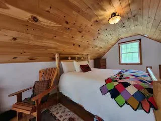 Cozy bedroom with sloped wooden ceiling, a bed with a colorful patchwork quilt, a wooden rocking chair, and a small window with natural light.