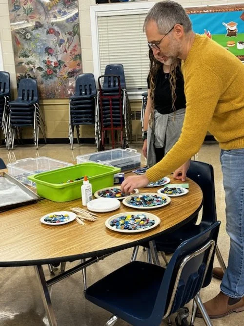 A man in a mustard yellow sweater and glasses is arranging colorful decorated cookies on plates at a round table. There are additional plates, a green container, a bottle of glue, and toothpicks on the table. In the background, a girl with long brown hair is partially visible, and there are stacked chairs and a colorful mural on the wall.